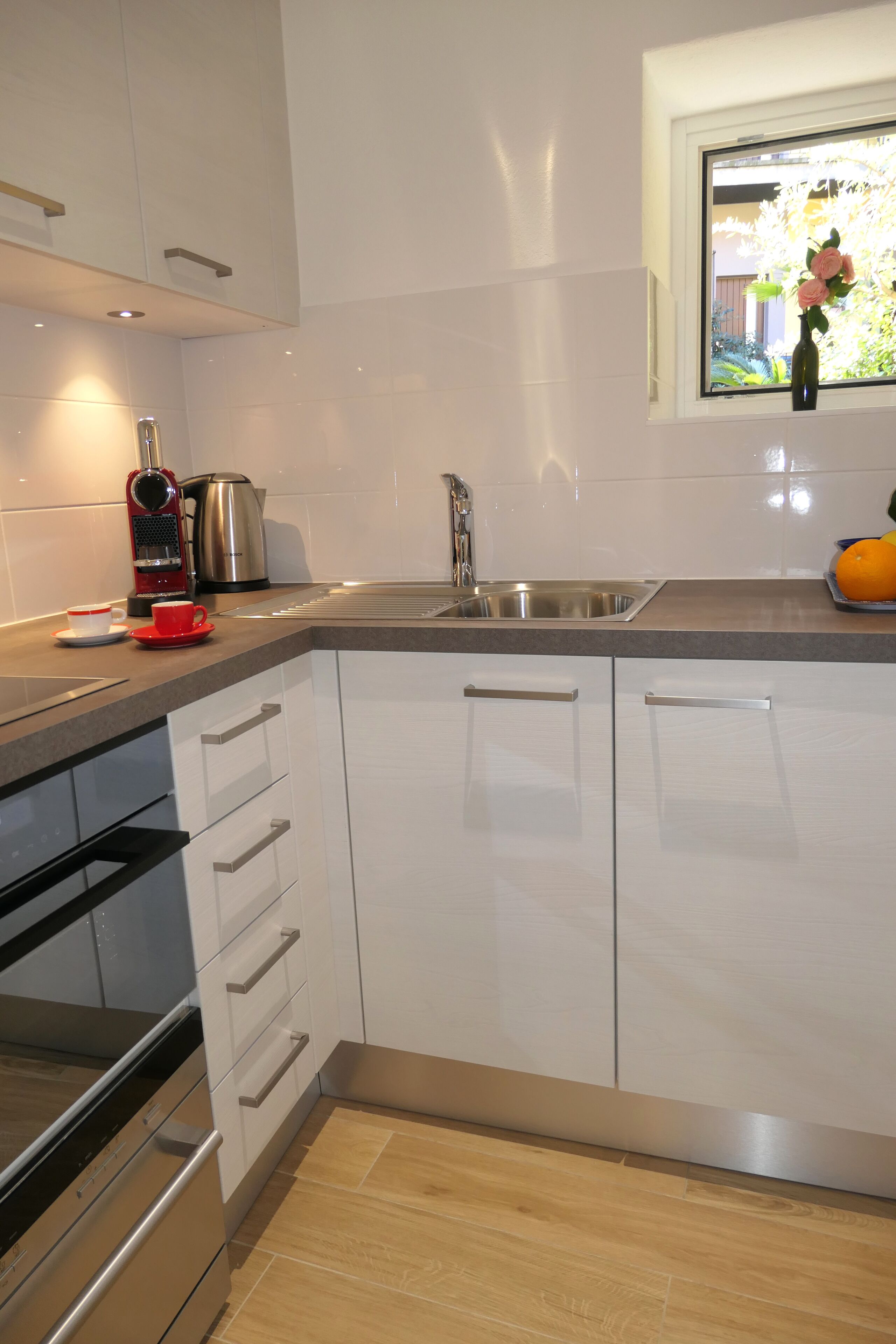 A modern kitchen with white cabinets, stainless steel appliances, and a window with a view of greenery.