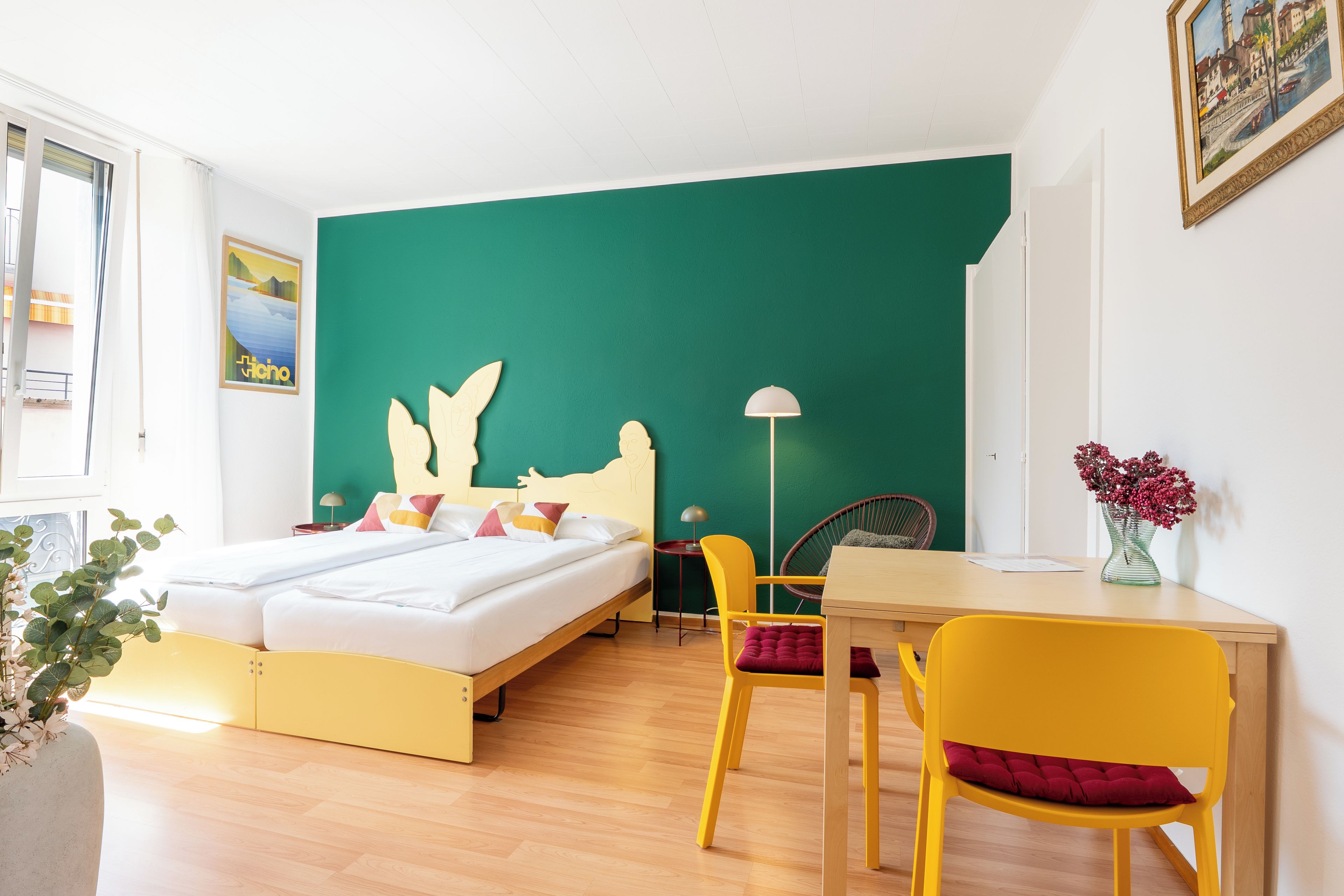 Una habitación con cama, escritorio, silla, lámpara y un jarrón con flores.