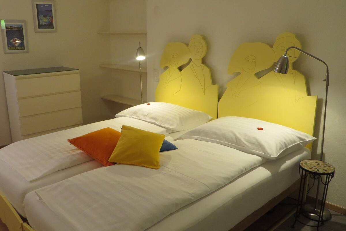 Una cama doble con ropa de cama blanca, dos almohadas (una naranja), una lámpara de noche y una mesita redonda.