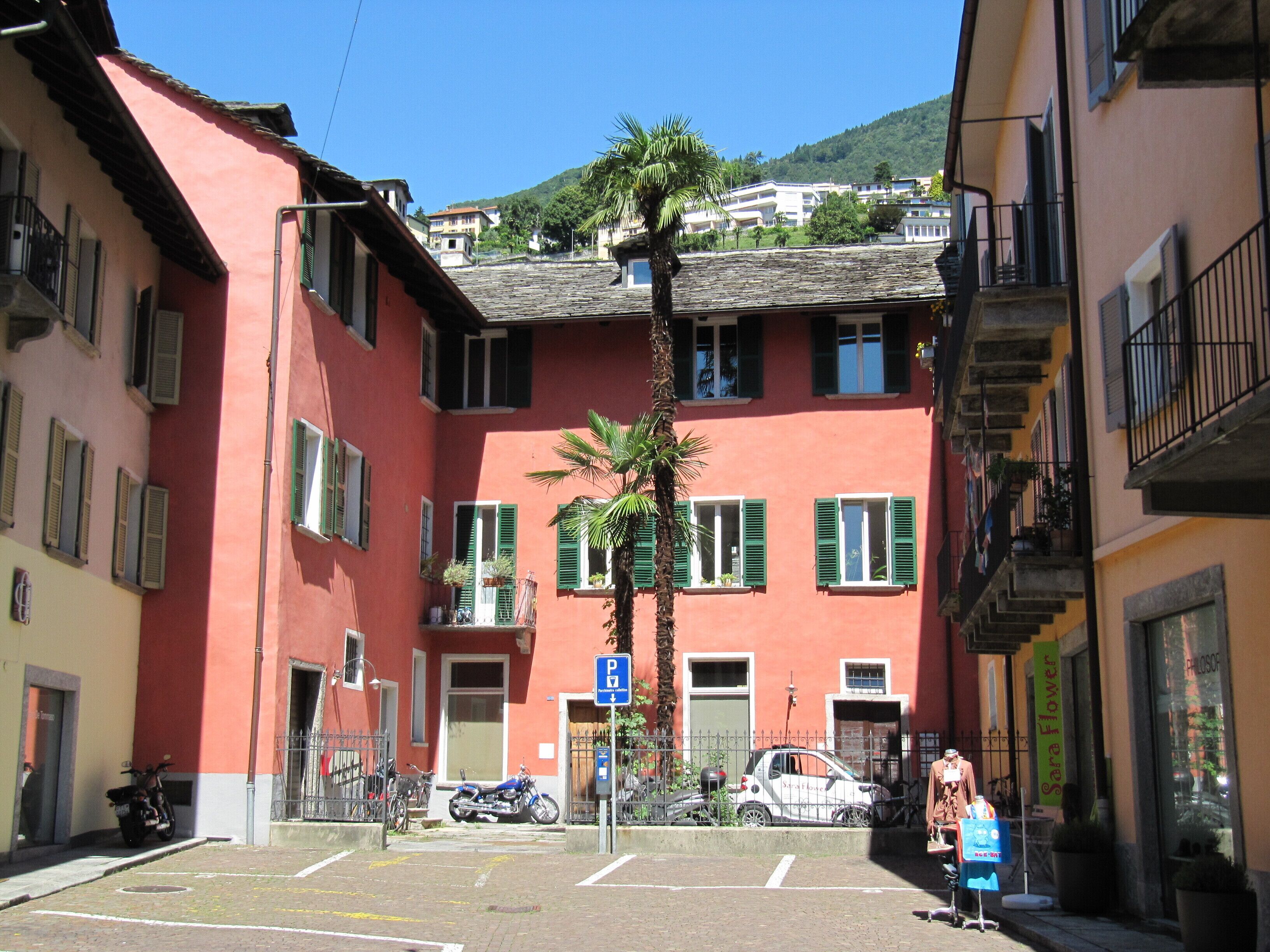 A multi-story building with a red facade, green shutters, and a central courtyard with a palm tree.