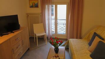 Hotelkamer met een televisie op een houten dressoir, een witte stoel, een bed met kussens, een vaas met rode bloemen en een raam met uitzicht op een balkon.