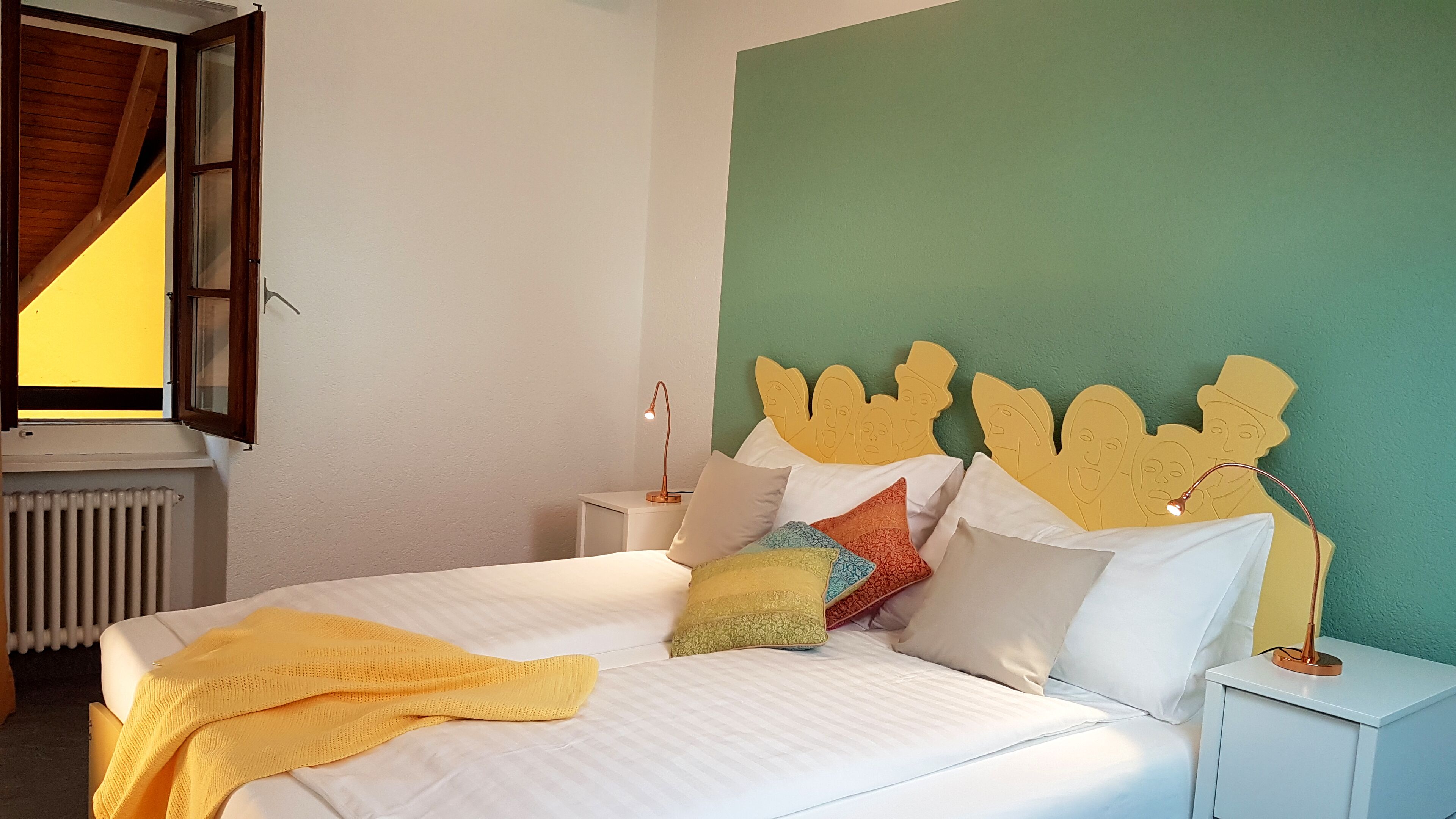 A neatly made bed with a yellow blanket, a headboard with face cutouts, and a bedside lamp.