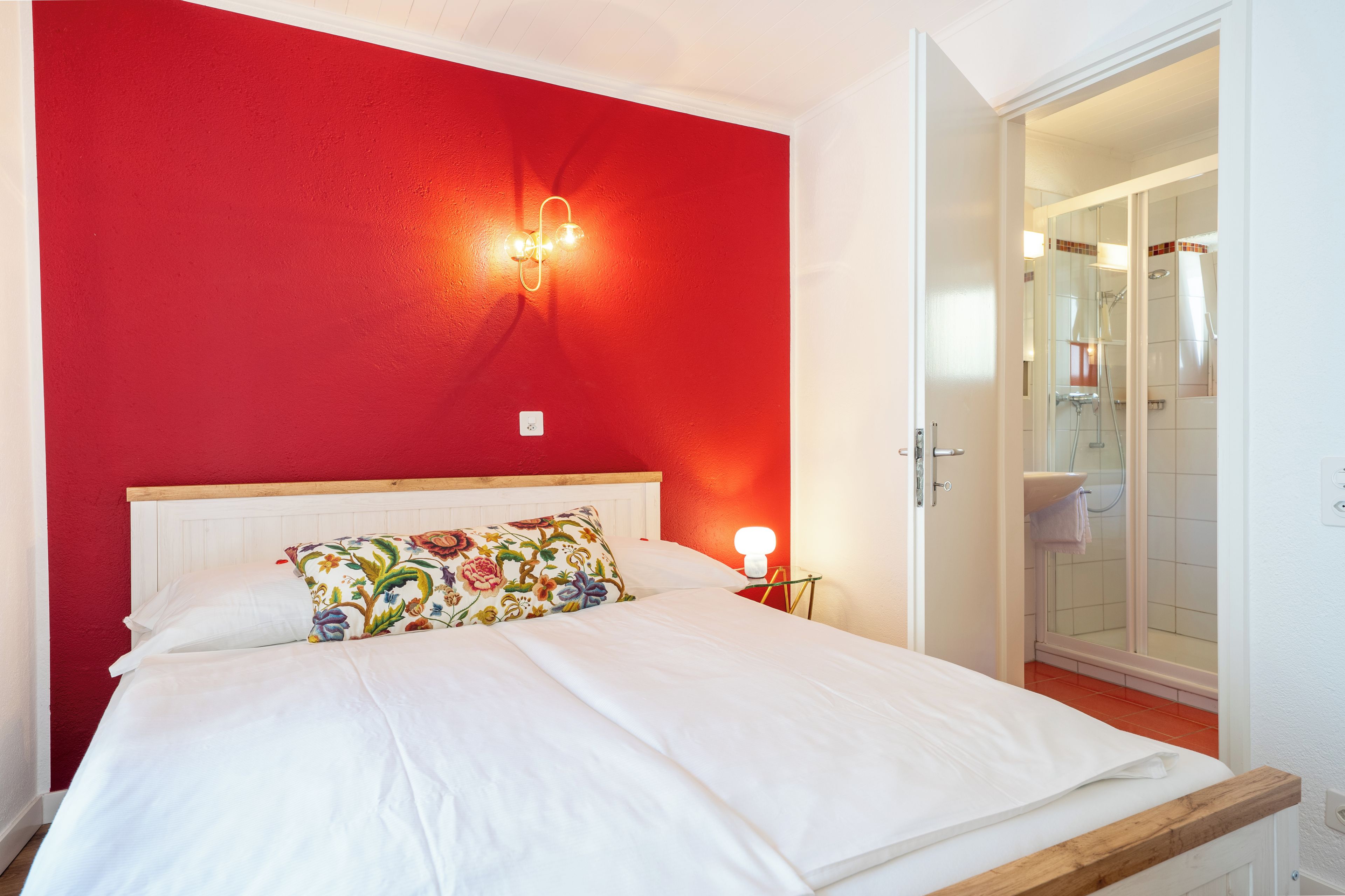 Un dormitorio con pared roja, cabecera de madera, cama blanca, almohada colorida, lámpara de noche y puerta abierta a un baño.