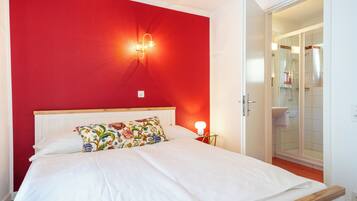 Un dormitorio con pared roja, cabecera de madera, cama blanca, almohada colorida, lámpara de noche y puerta abierta a un baño.