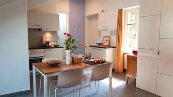 A compact kitchen with white cabinets, a wooden dining table, and two chairs. A vase with flowers is on the table.