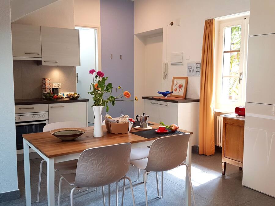 A compact kitchen with white cabinets, a wooden dining table, and two chairs. A vase with flowers is on the table.