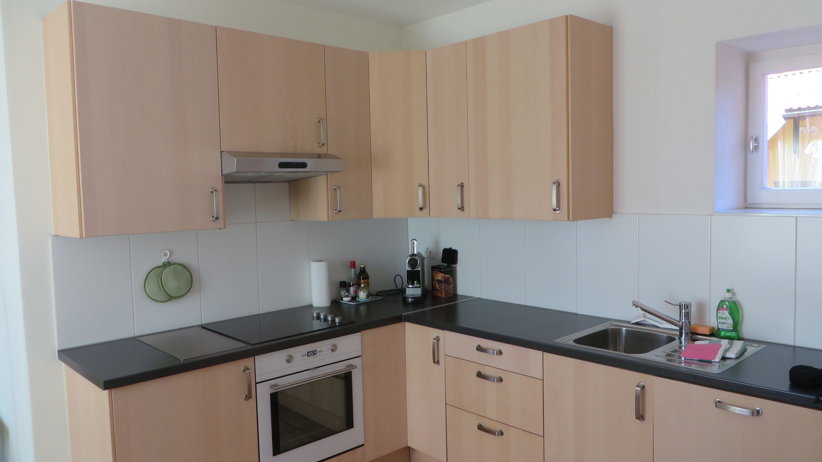 A kitchen with light wood cabinets, a black countertop, a stainless steel sink, and a window with a view.