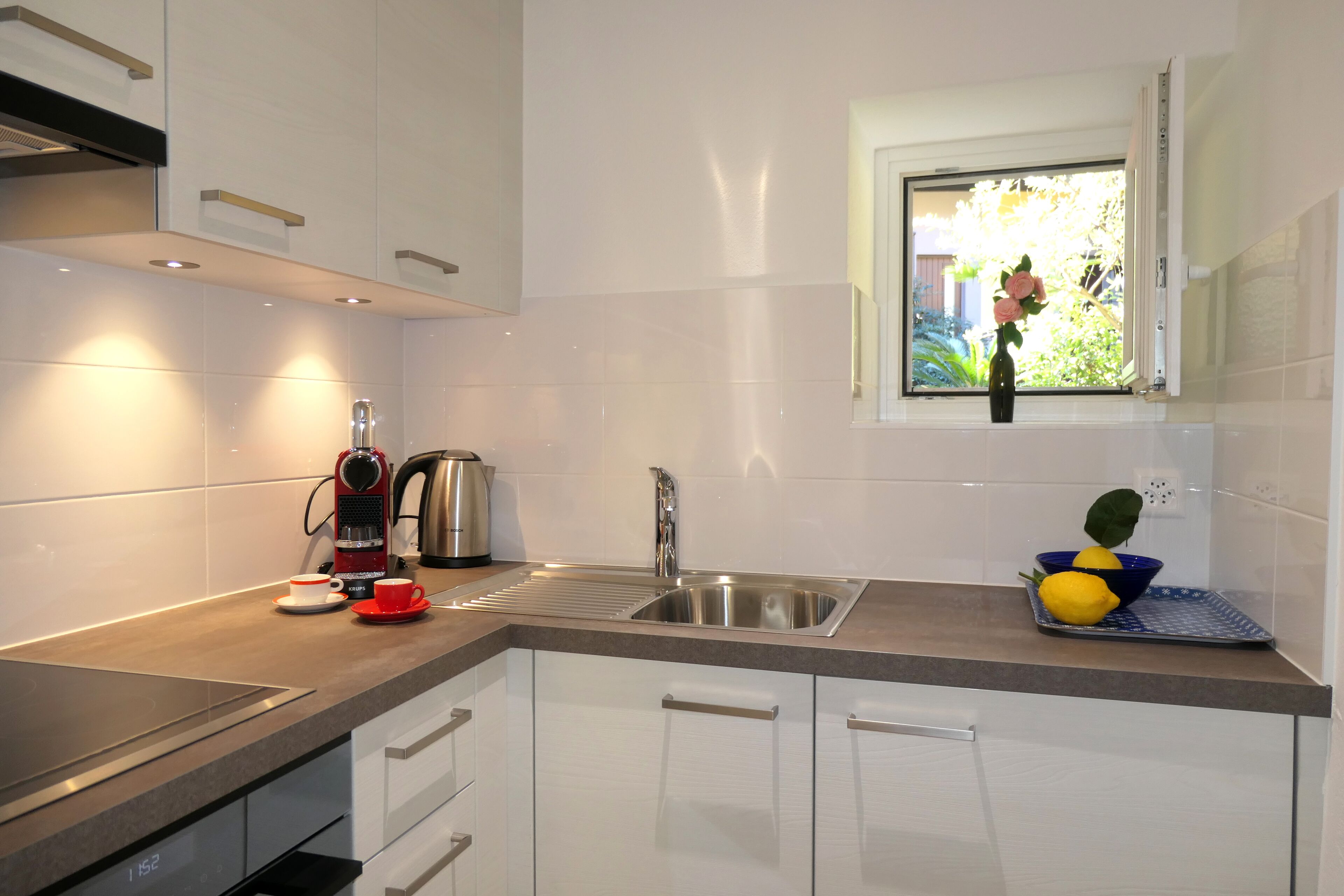 A modern kitchen with white cabinets, a stainless steel sink, a kettle, and a window with a view of greenery.
