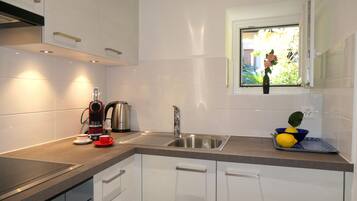A modern kitchen with white cabinets, a stainless steel sink, a kettle, and a window with a view of greenery.