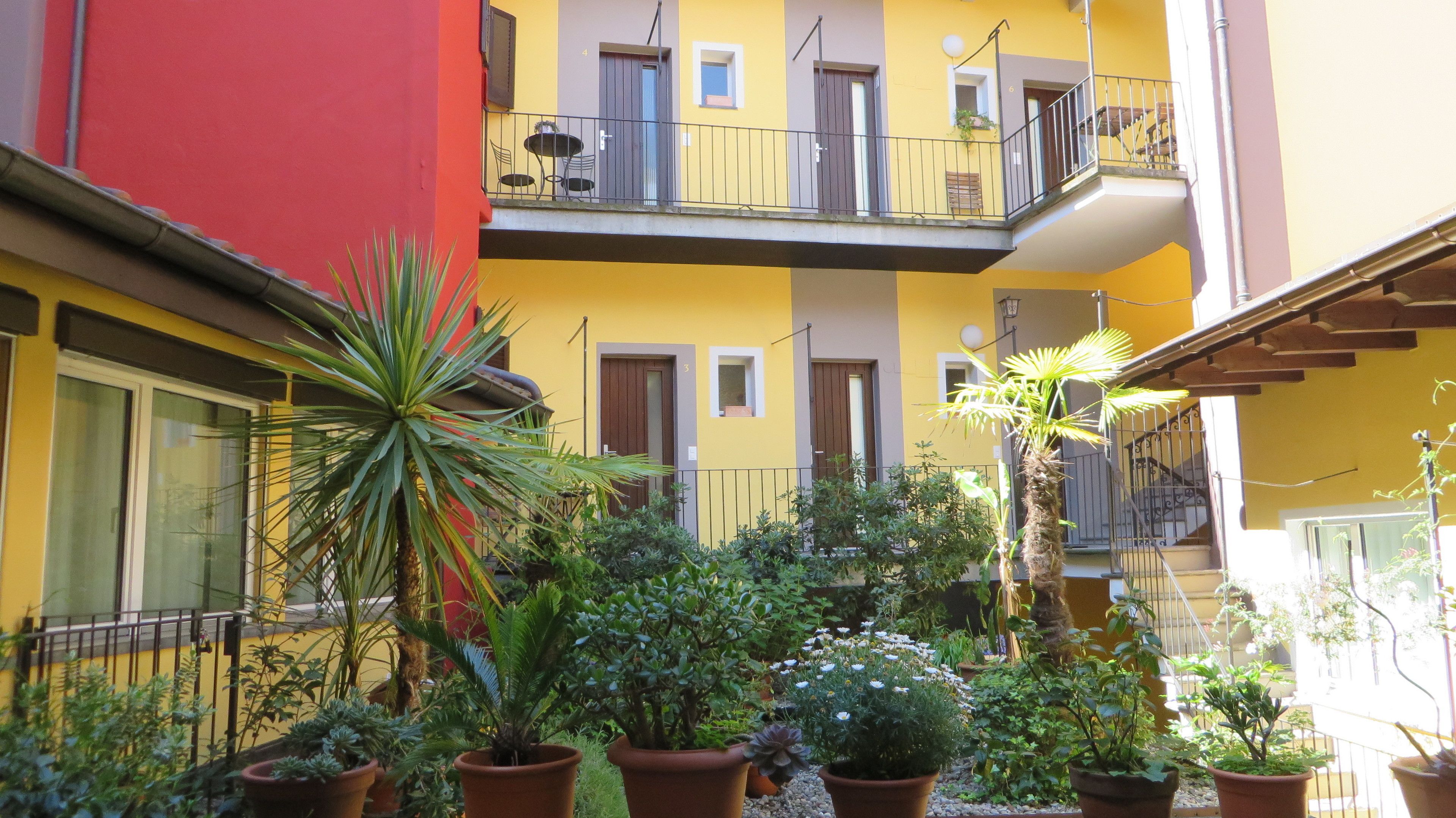 Um pátio com plantas em vasos, um edifício vermelho e um edifício amarelo com sacadas.