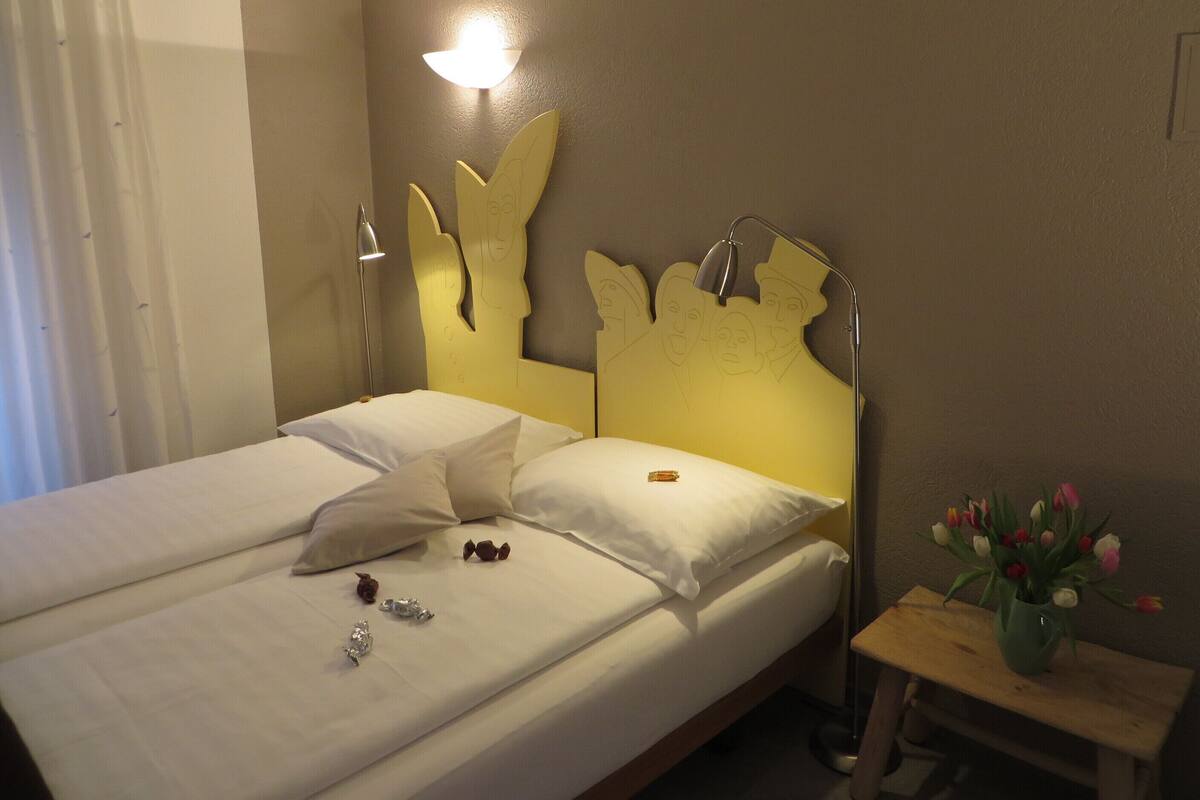 Una cama bien hecha con almohadas, una lámpara de noche y una pequeña mesita de madera con un jarrón de flores.