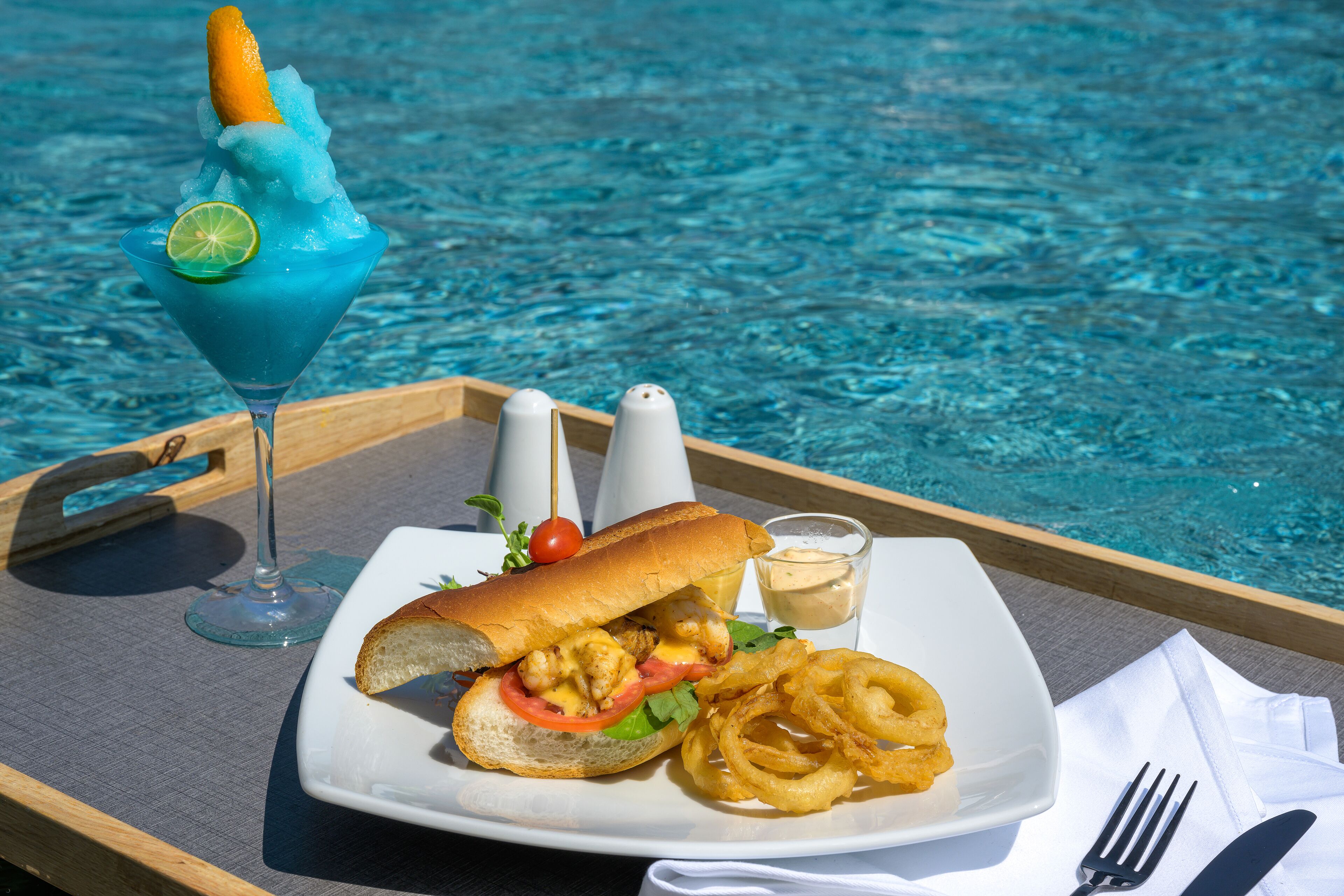 A tray with a sandwich, onion rings, a blue drink, and condiments by a pool.