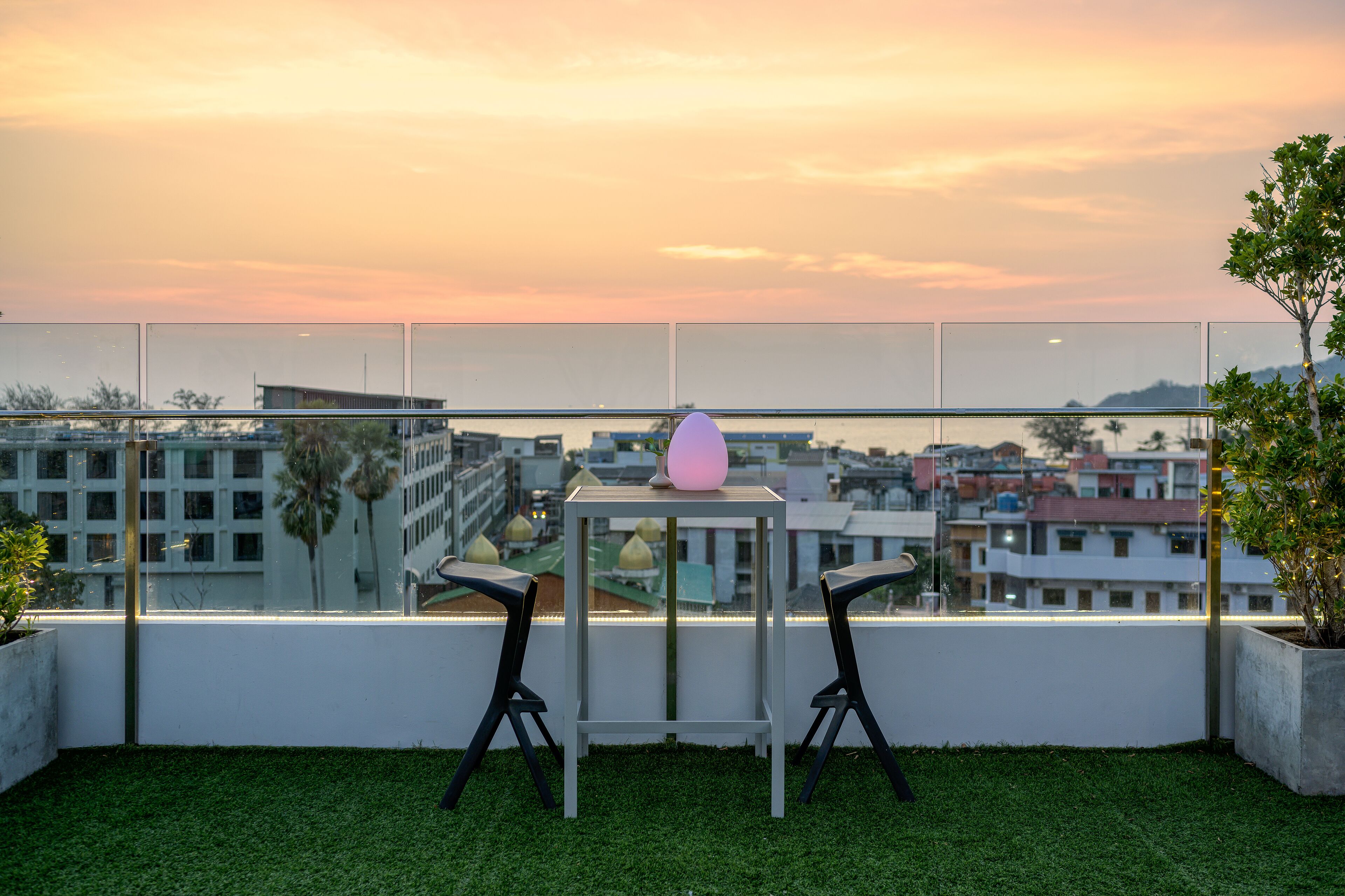 A balcony with two chairs and a table, overlooking a cityscape at sunset.