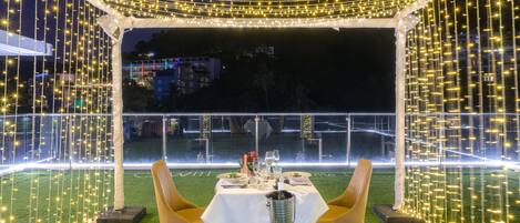 An outdoor dining area with a table set for two, surrounded by greenery and string lights.