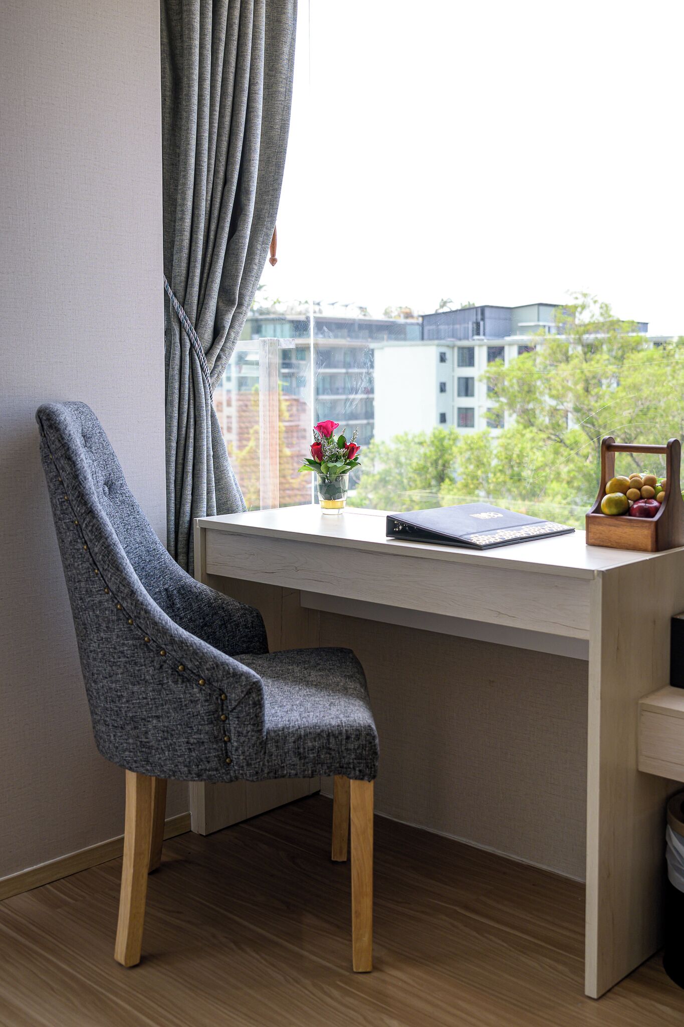 A room with a desk, a chair, a window with curtains, a vase with flowers, and a fruit basket.