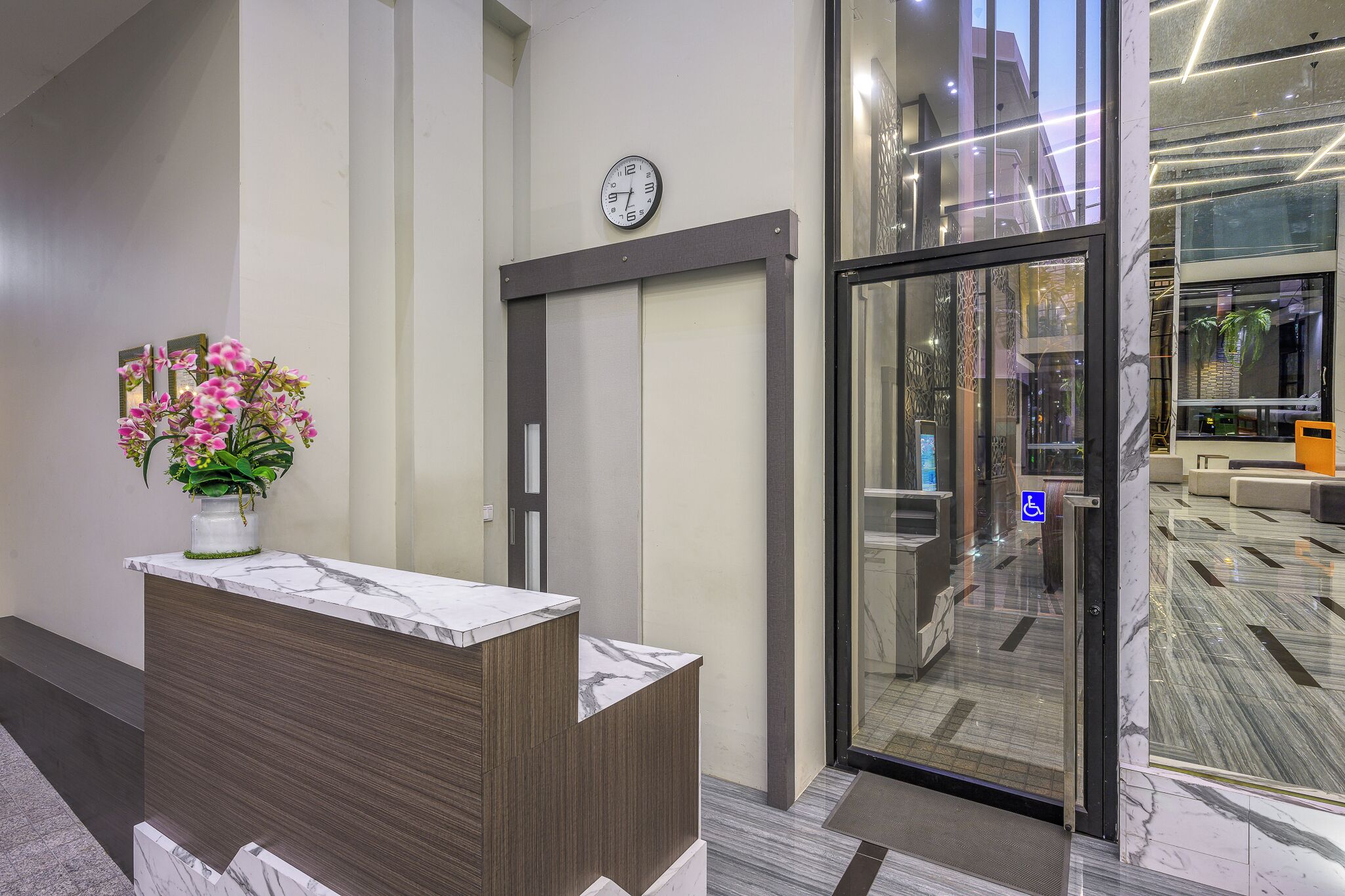 A reception area with a marble counter, a clock, and a glass door leading to a lobby with a patterned floor.