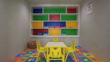 Uma área de recreação infantil com uma mesa branca e cadeiras amarelas, cercada por prateleiras coloridas e um tapete de atividades.