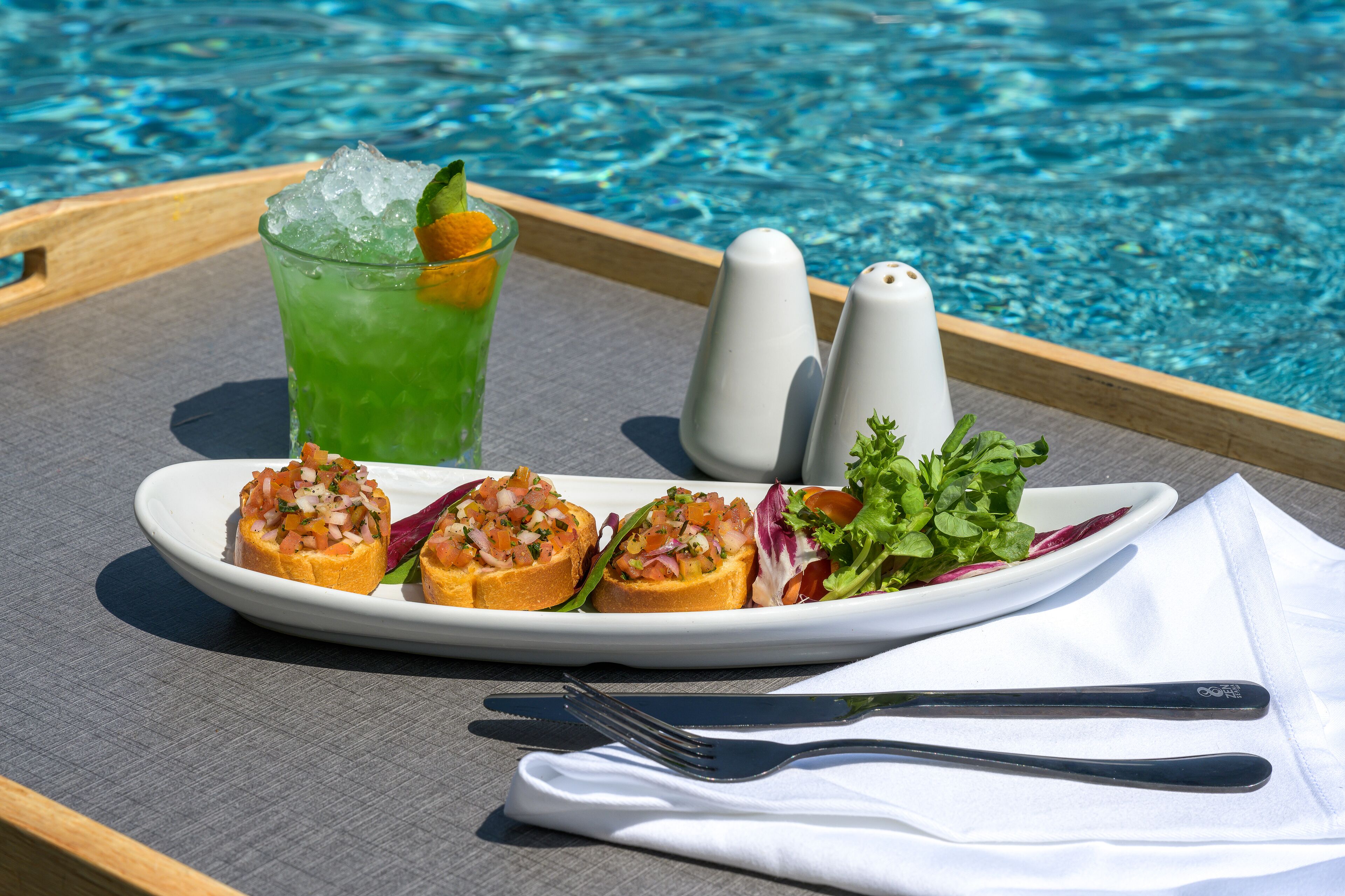 Una bandeja con una bebida verde, un plato de aperitivos y saleros y pimenteros junto a una piscina.