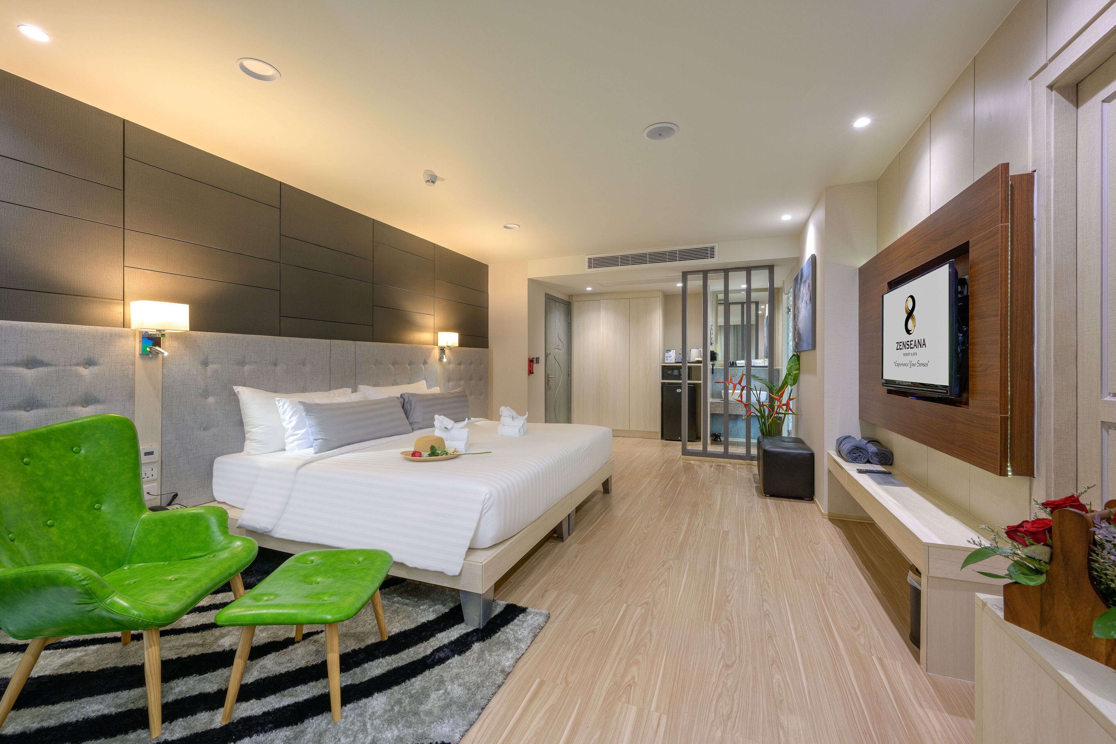 A modern hotel room with a bed, green chairs, a black and white rug, a TV, and a reception desk with a plant.