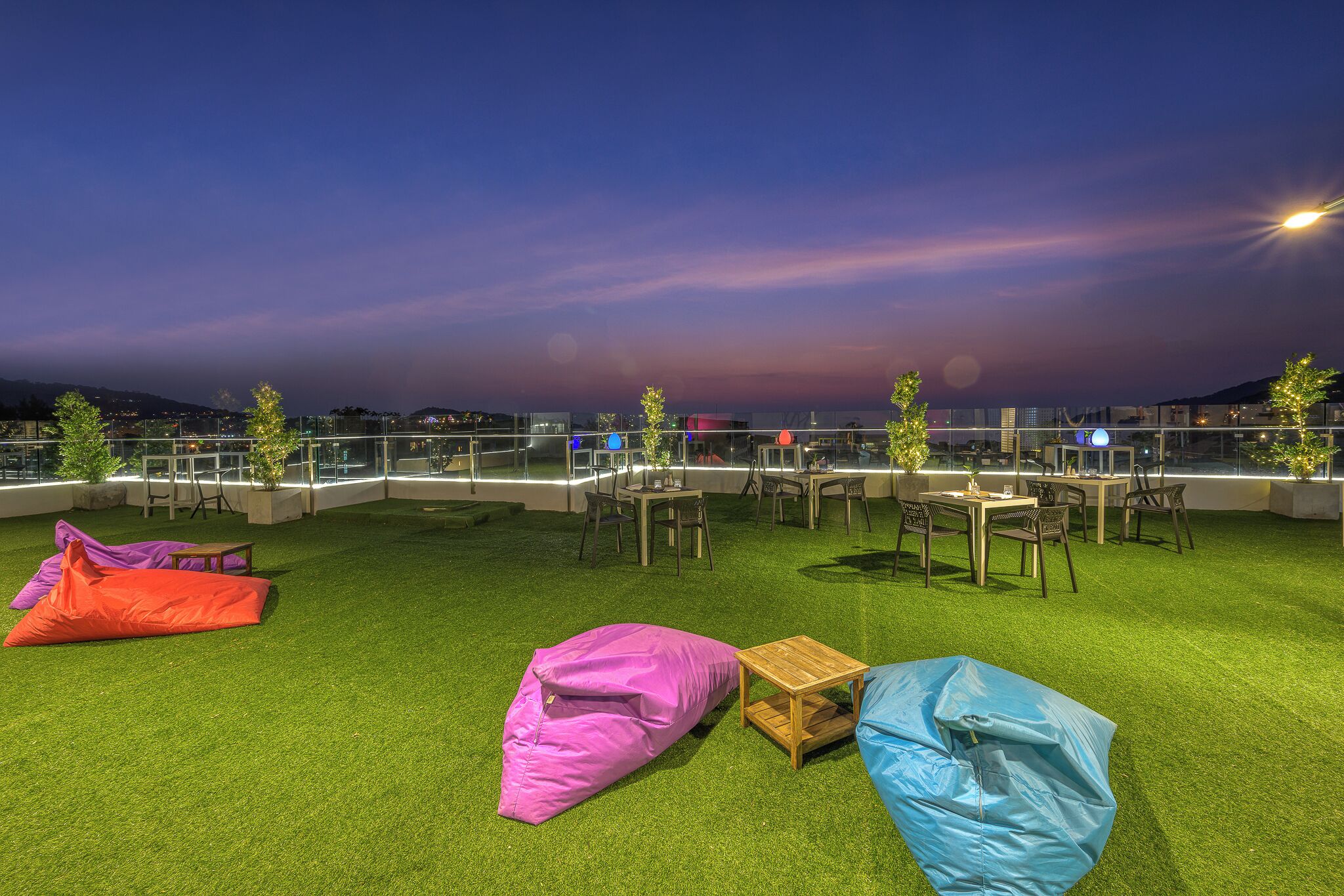 An outdoor seating area with tables and chairs, surrounded by a glass railing, and a view of the cityscape at dusk.