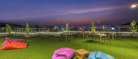 An outdoor seating area with tables and chairs, surrounded by a glass railing, and a view of the cityscape at dusk.