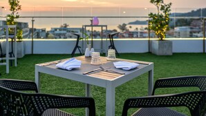 Un área de comedor en la azotea con una mesa preparada para dos personas, rodeada de sillas, y vista a la ciudad al atardecer.