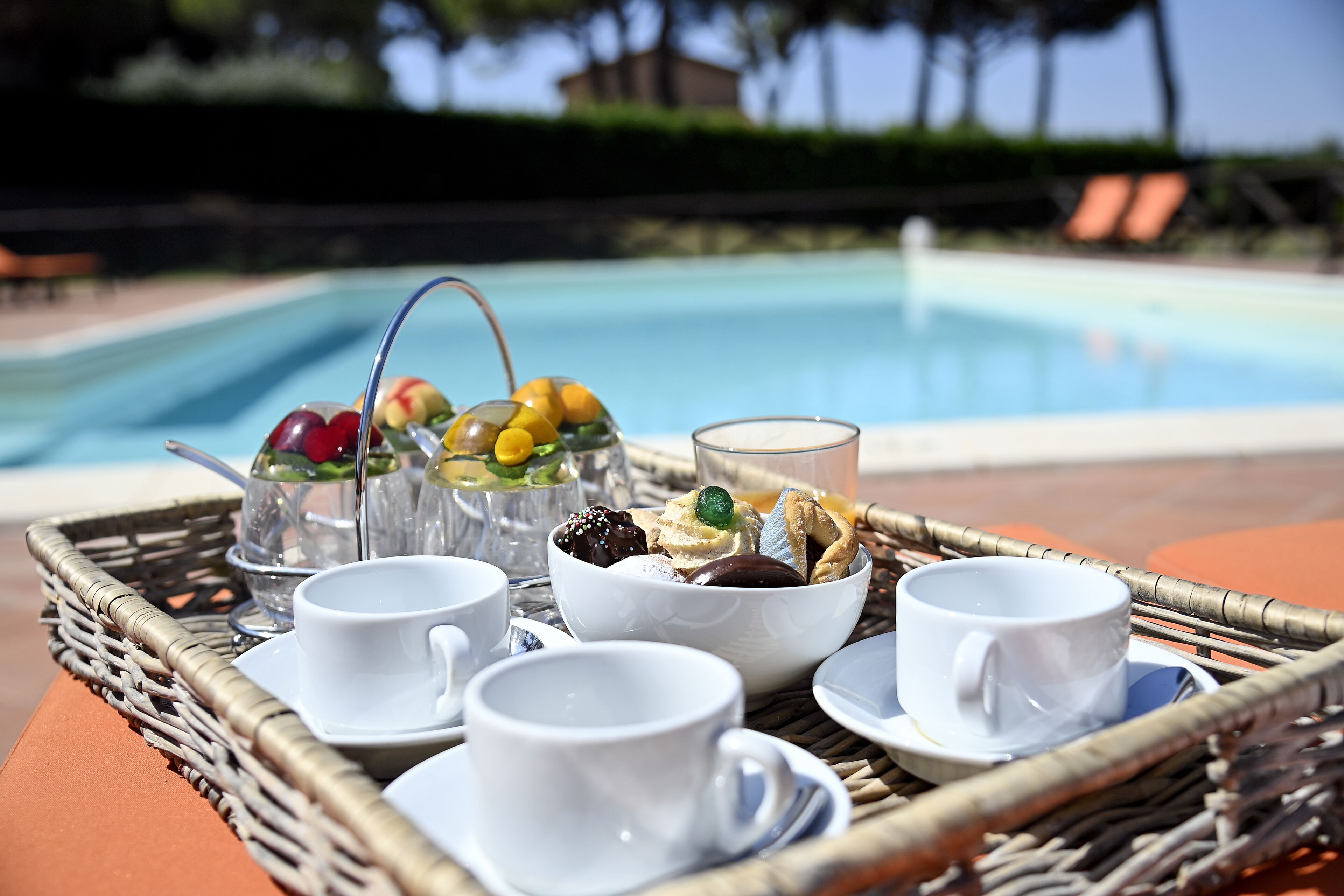 A tray with white cups, a glass, and a bowl of assorted fruits and pastries by a pool.