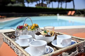 Una bandeja con tazas blancas, un vaso y un bol de frutas y pasteles junto a una piscina.