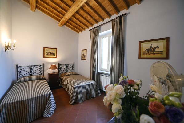 Una habitación con dos camas, techo de madera, ventana con cortinas, ventilador de pared y un cuadro enmarcado.
