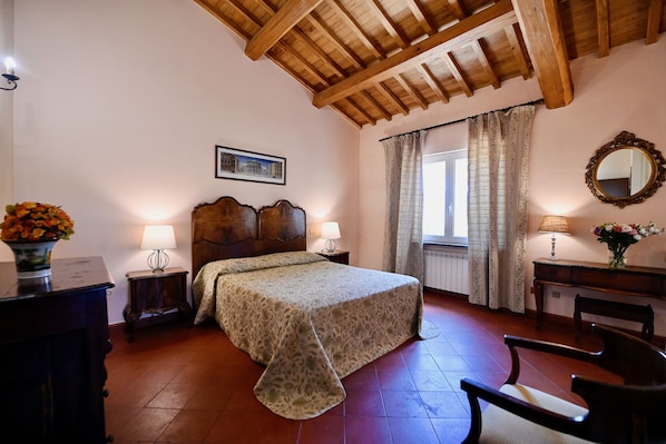 Un dormitorio con techo de madera, una cama con colcha estampada, dos lámparas de noche, una mesita de noche, un aparador, una silla, una ventana con cortinas y un espejo en la pared.