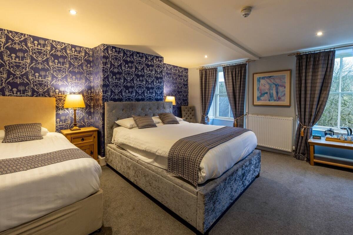 A hotel room with two beds, a large window with curtains, a radiator, and a small table with a teapot.