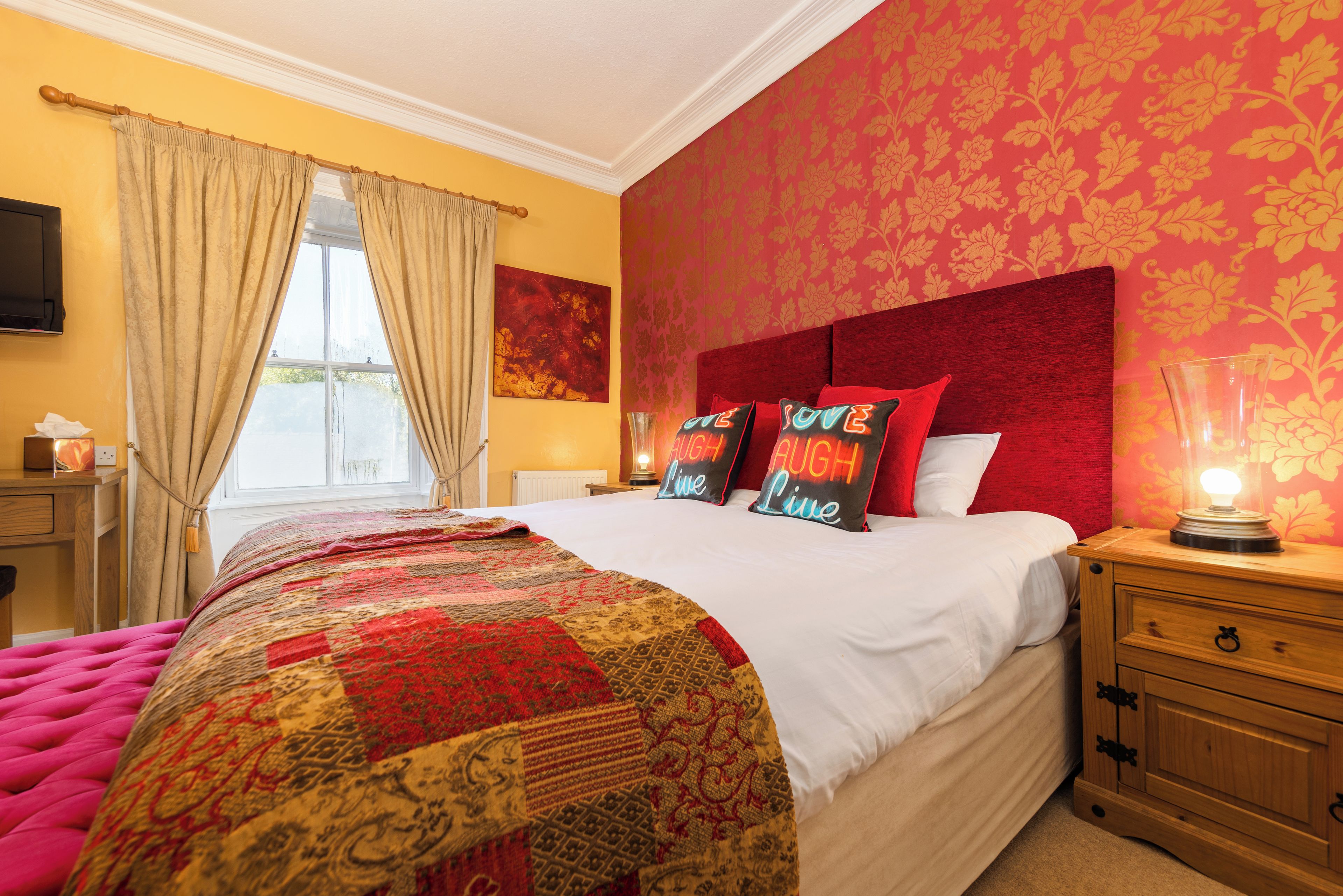Habitación de hotel con una cama grande, una mesita de noche con lámpara, una ventana con cortinas y una pared con estampado floral.