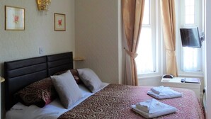 A hotel room with a bed, pillows, a TV, and a window with curtains. - The Winchester (Great Yarmouth)