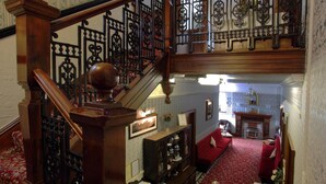 A grand staircase with ornate railings leading to a second floor. A red carpet runs along the floor, and a fireplace is visible in the background. - The Winchester (Great Yarmouth)