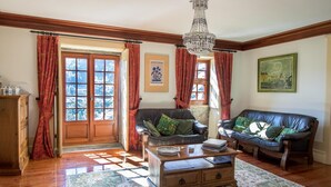 A living room with a wooden coffee table, two leather sofas, a chandelier, and a view through a large window with red curtains. - Casa do Monte (Baiao)