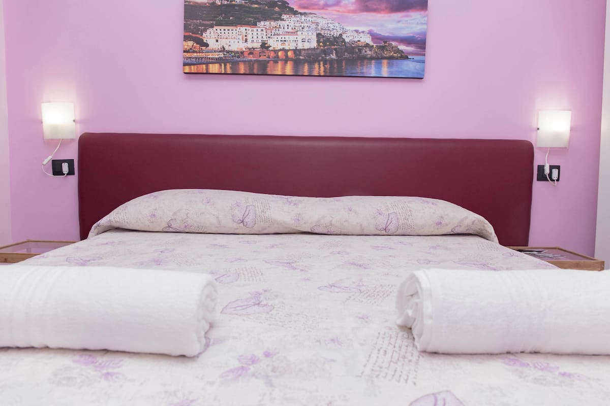 Una cama con cabecera roja, ropa de cama blanca y estampado floral morado. Hay dos lámparas de noche y un cuadro de un pueblo costero en la pared.