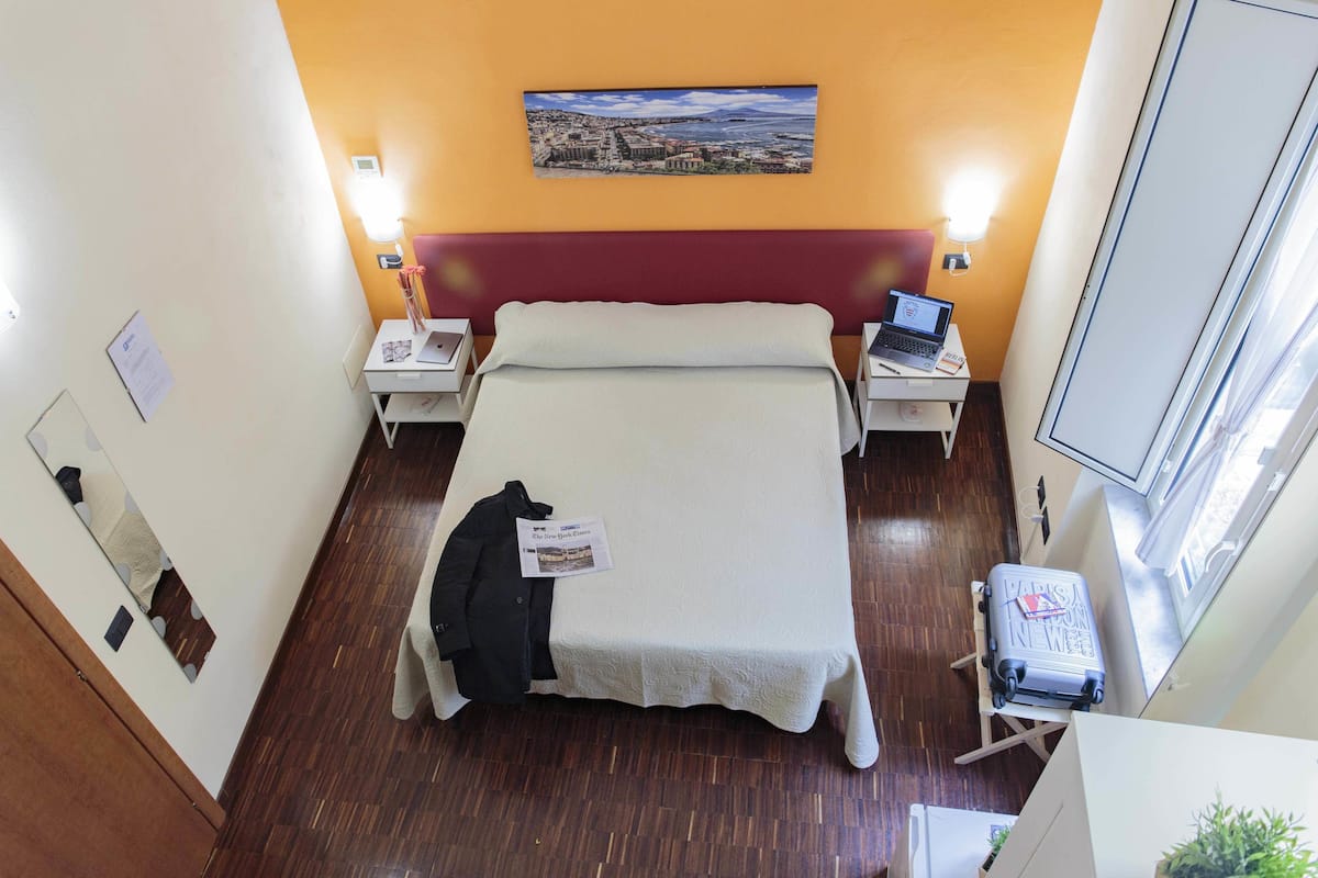 Una habitación de hotel con una cama, dos mesitas de noche, una computadora portátil, una maleta y una ventana.