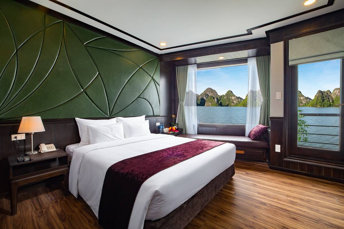 A large bed with a dark headboard, a bedside table with a lamp, and a view of a lake through a window with curtains.