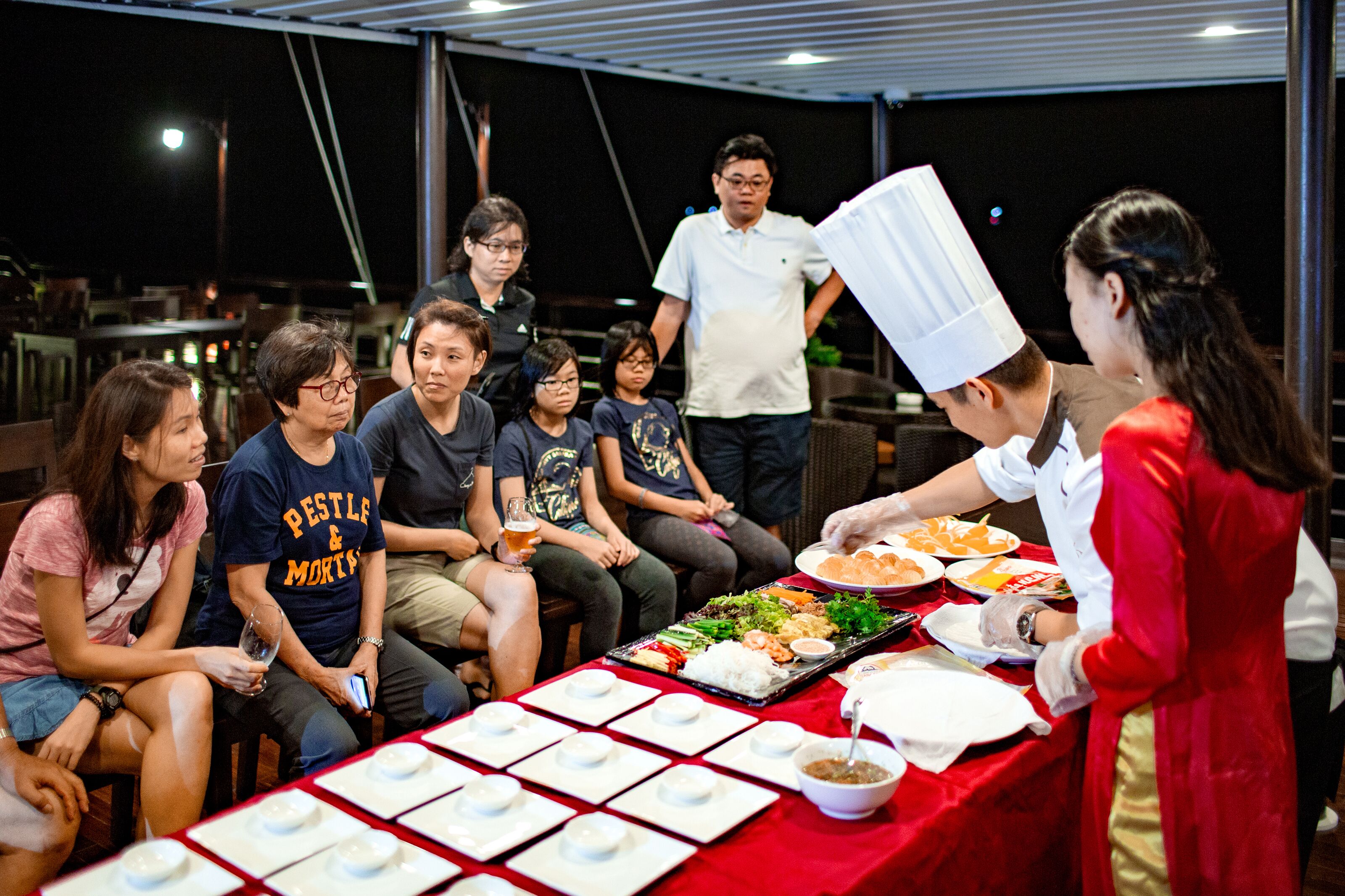 A group of people seated around a table with food and drinks, a chef serving food, and a tented outdoor setting.