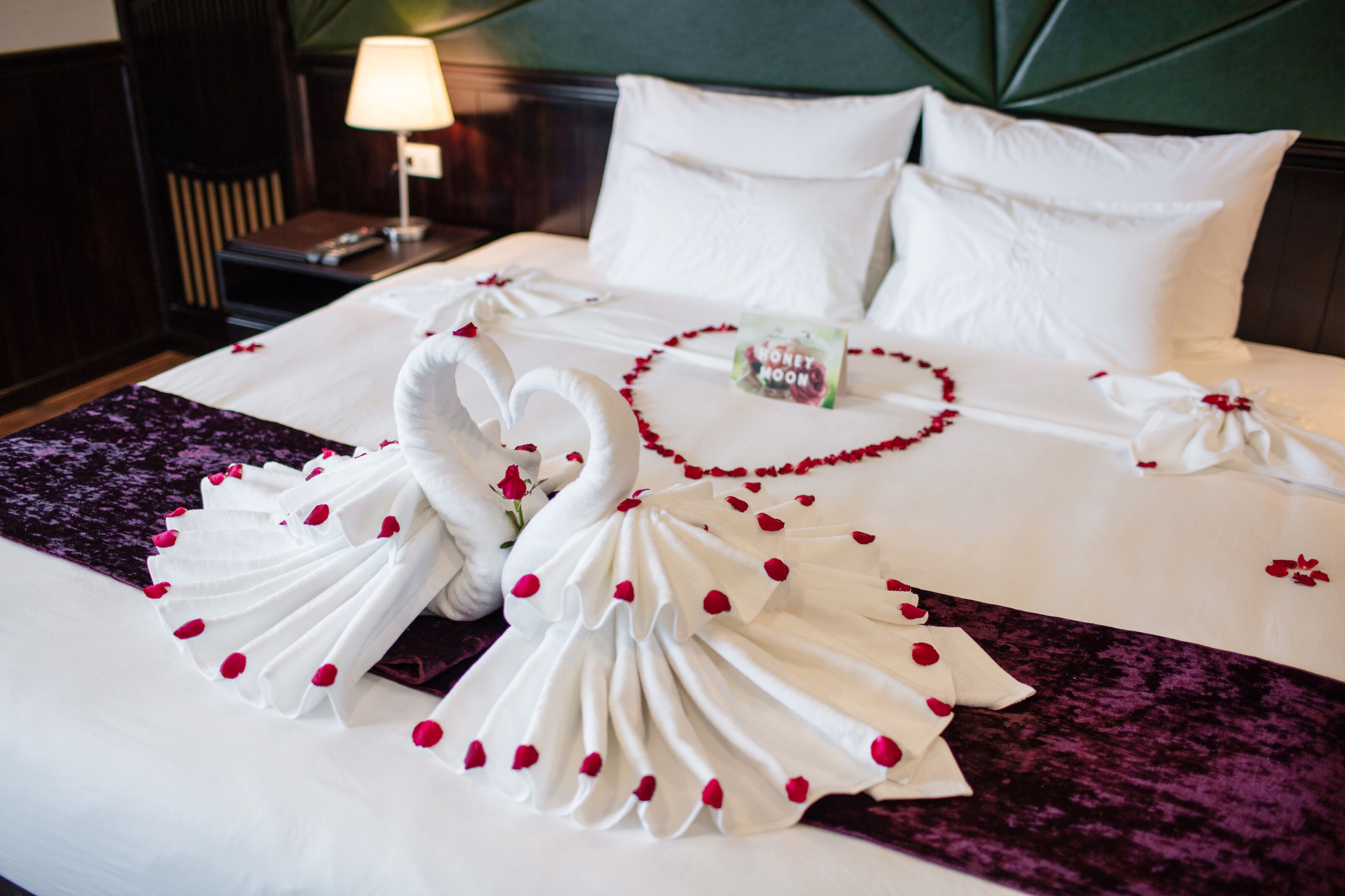 Un lit parfaitement fait, avec du linge de lit blanc, une composition en forme de cœur faite de pétales de roses et deux décorations en forme de cygne.
