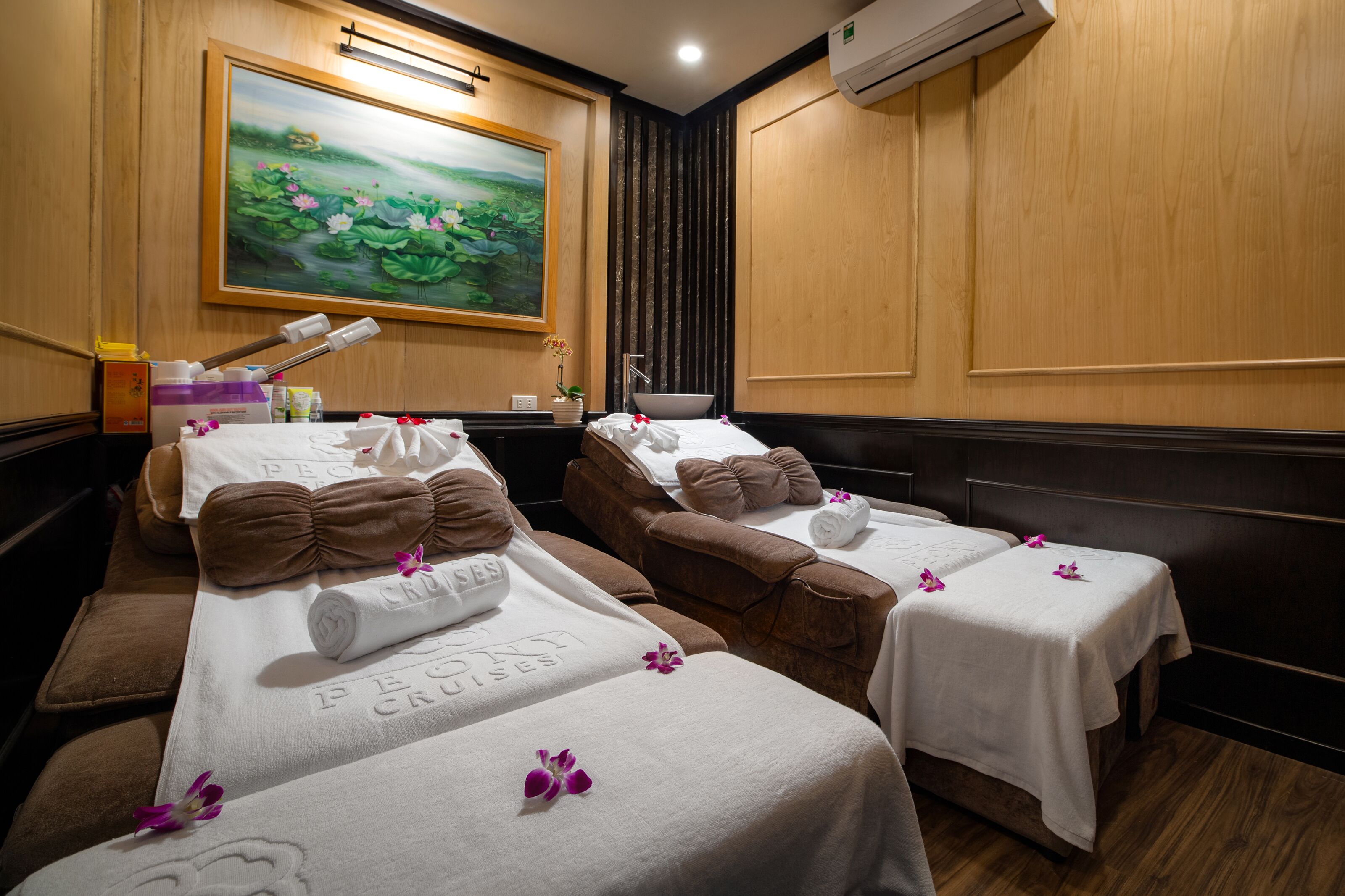 A room with two massage tables, each with a towel and a pillow. There is a painting on the wall and an air conditioning unit above the tables.