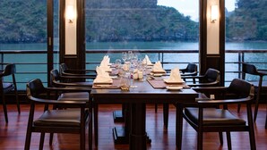 Mesa para seis personas preparada, con sillas, ubicada cerca de un ventanal con vistas a un cuerpo de agua y un acantilado.