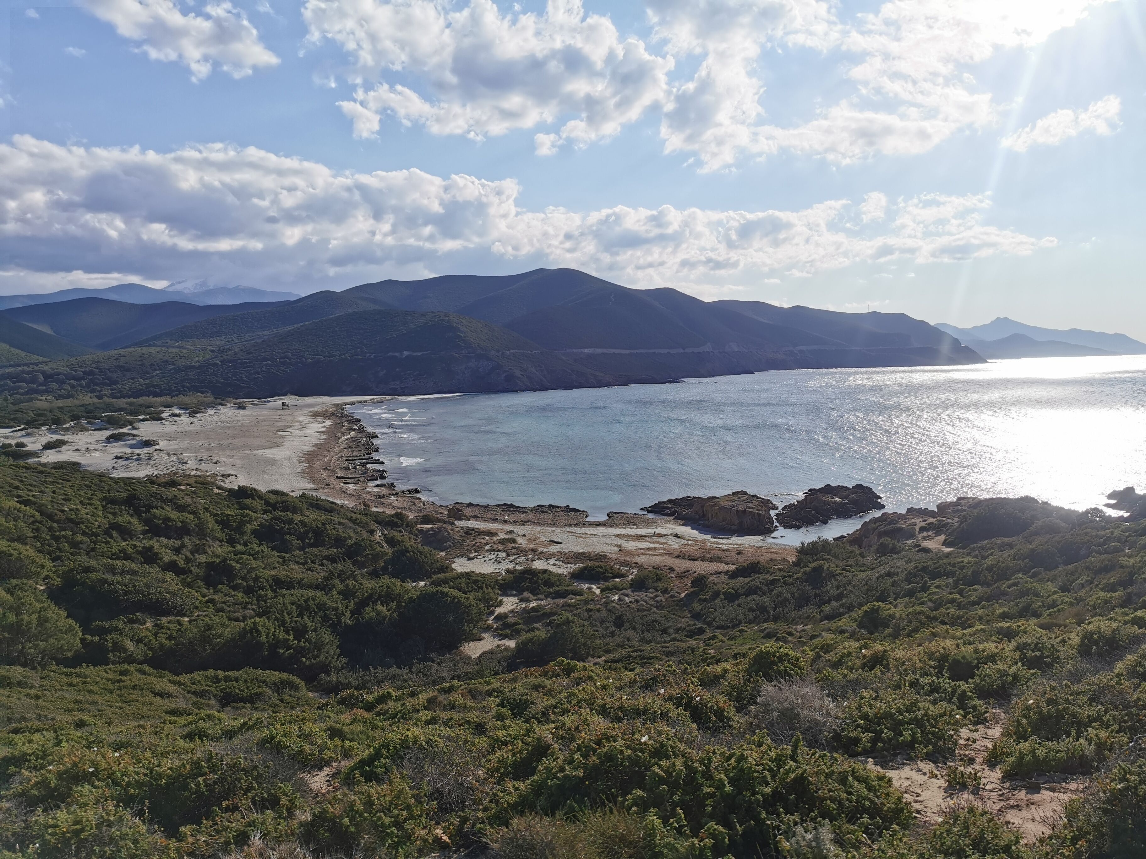 A coastal landscape with a beach, rocky outcrops, and distant mountains under a partly cloudy sky.