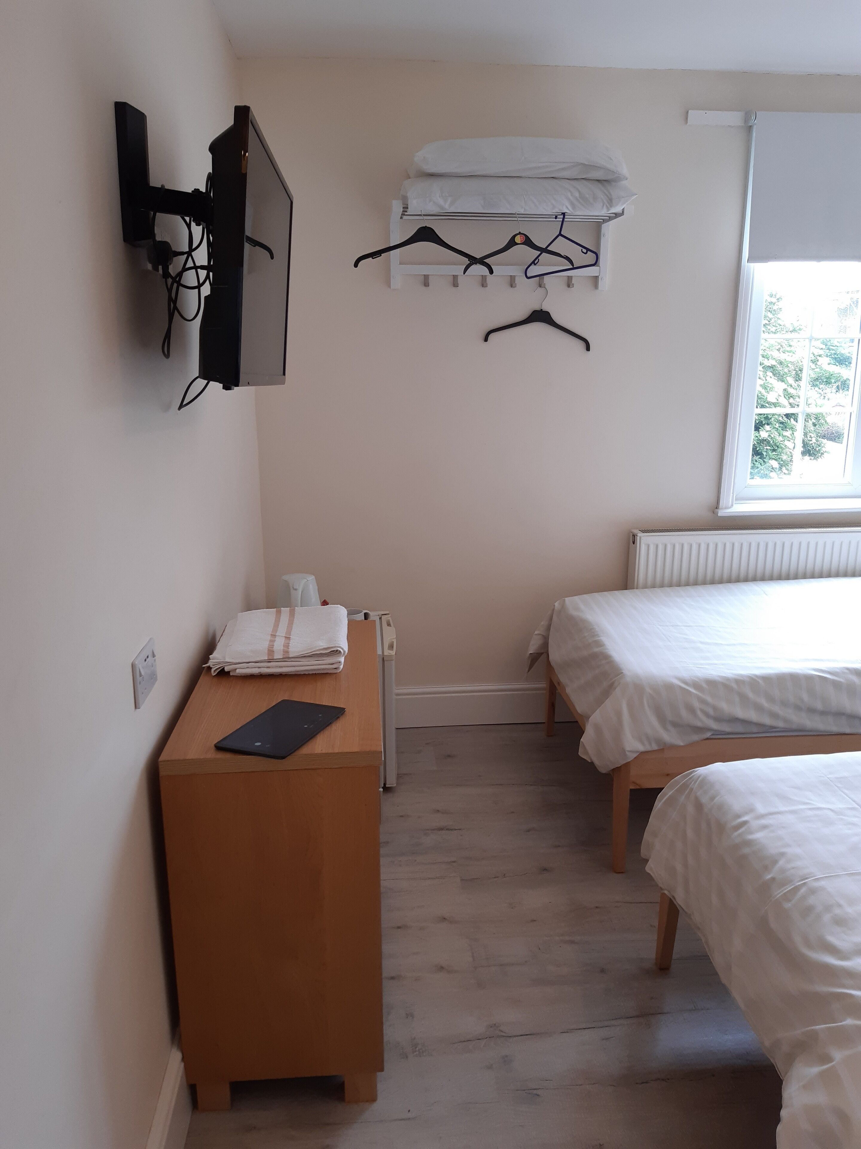 A bedroom with two beds, a TV mounted on the wall, a radiator, and a window with a view of trees.