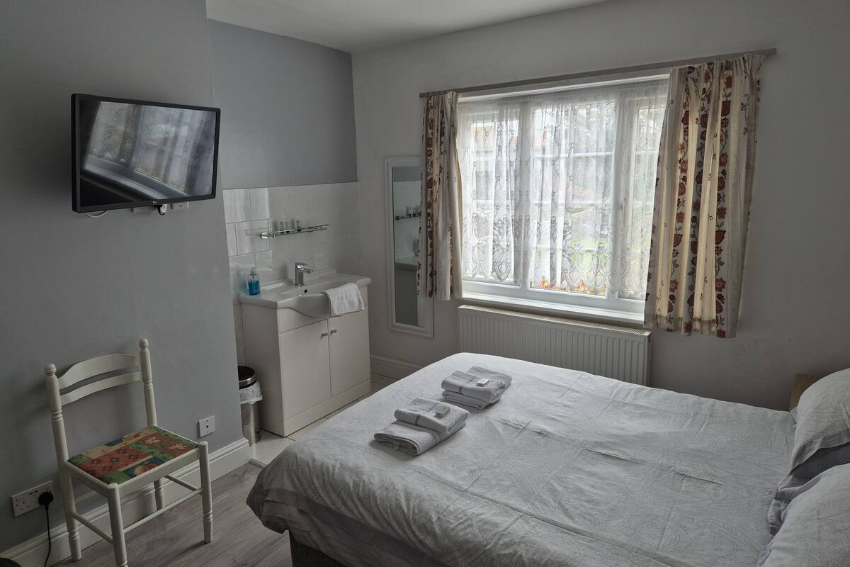 A bedroom with a bed, a chair, a TV mounted on the wall, a window with curtains, and a small bathroom with a sink and mirror.