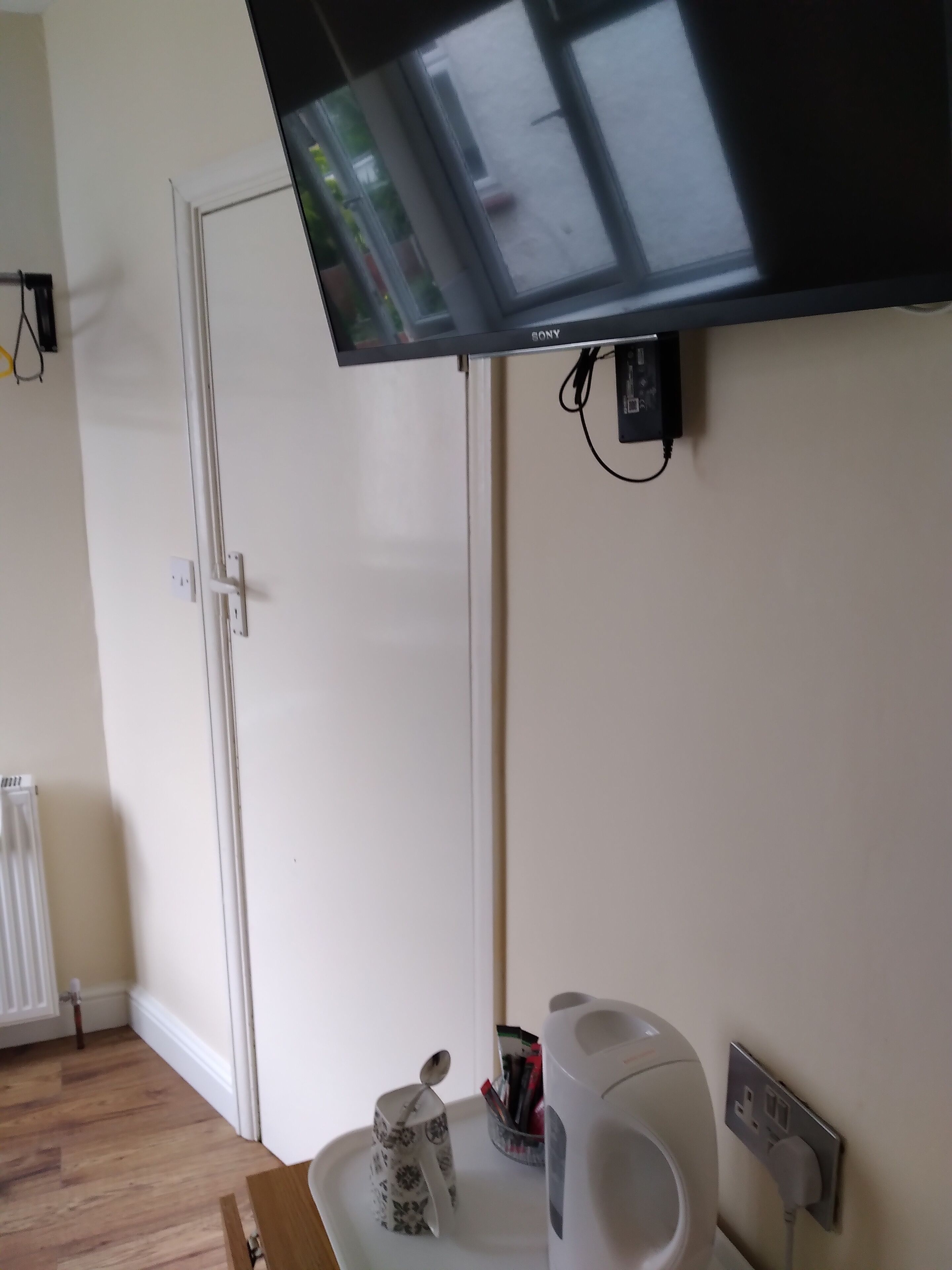 Een kamer met een wandgemonteerde flatscreen televisie, een witte radiator, een houten vloer, een witte deur en een dienblad met een mok en een waterkoker.