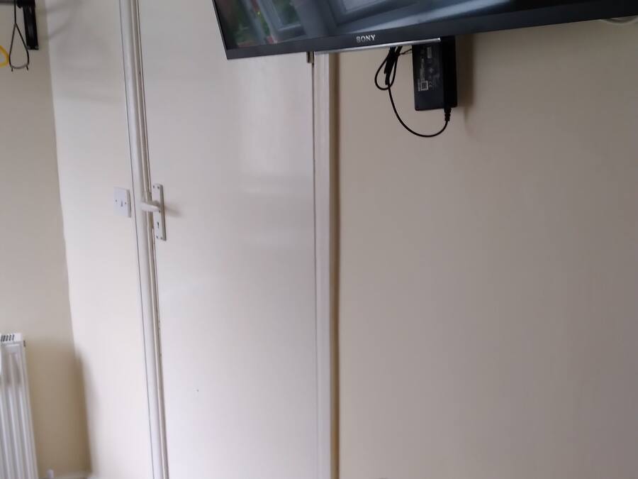 A room with a flat-screen TV mounted on the wall, a white radiator, a wooden floor, a white door, and a tray with a mug and a kettle.