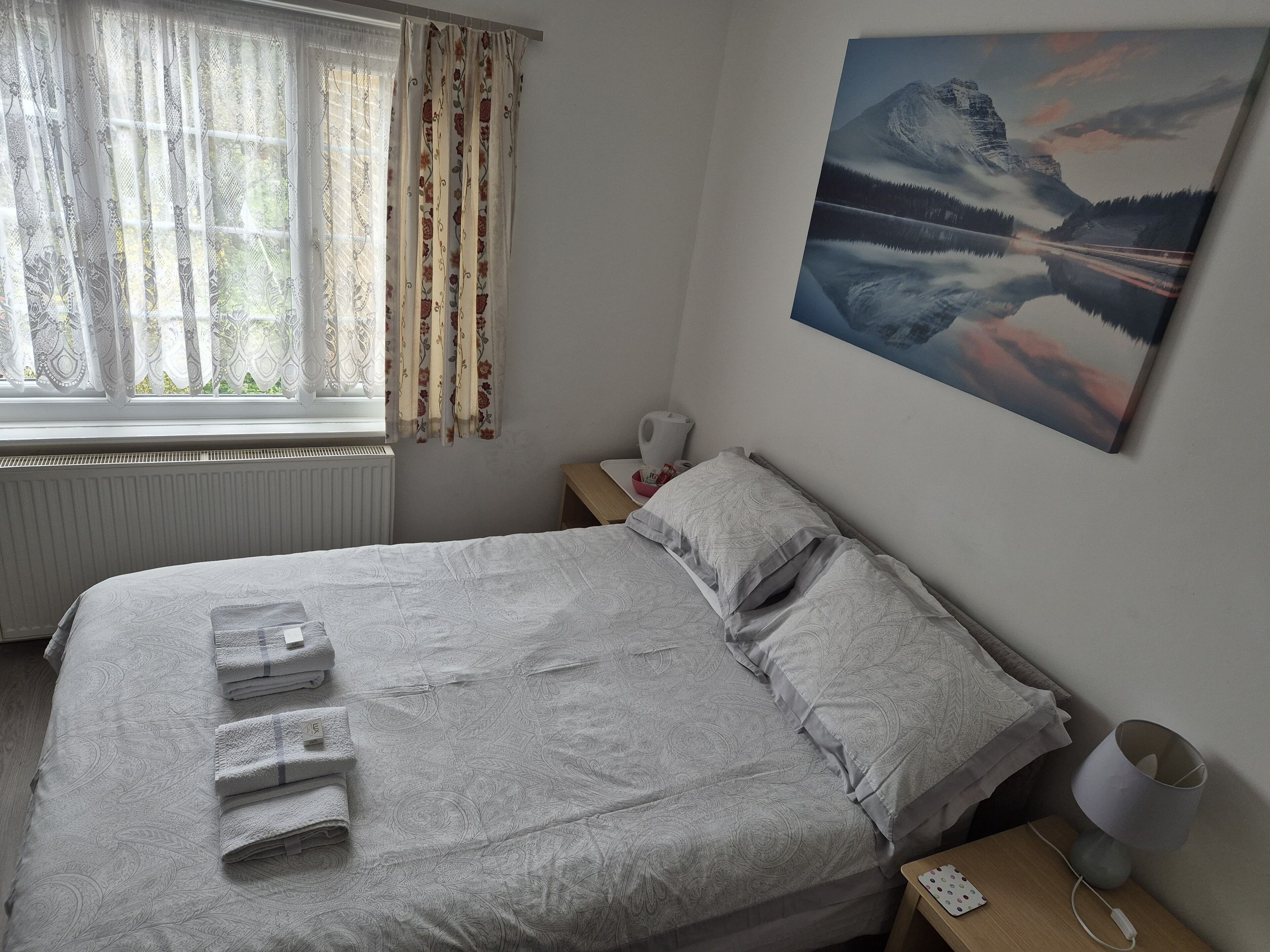 Een netjes opgemaakt bed met handdoeken, een nachtkastje met een lamp, een schilderij van een berglandschap en een raam met gordijnen.