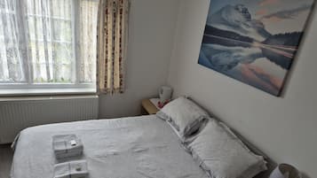 A neatly made bed with folded towels, a bedside table with a lamp, a painting of a mountain landscape, and a window with curtains.