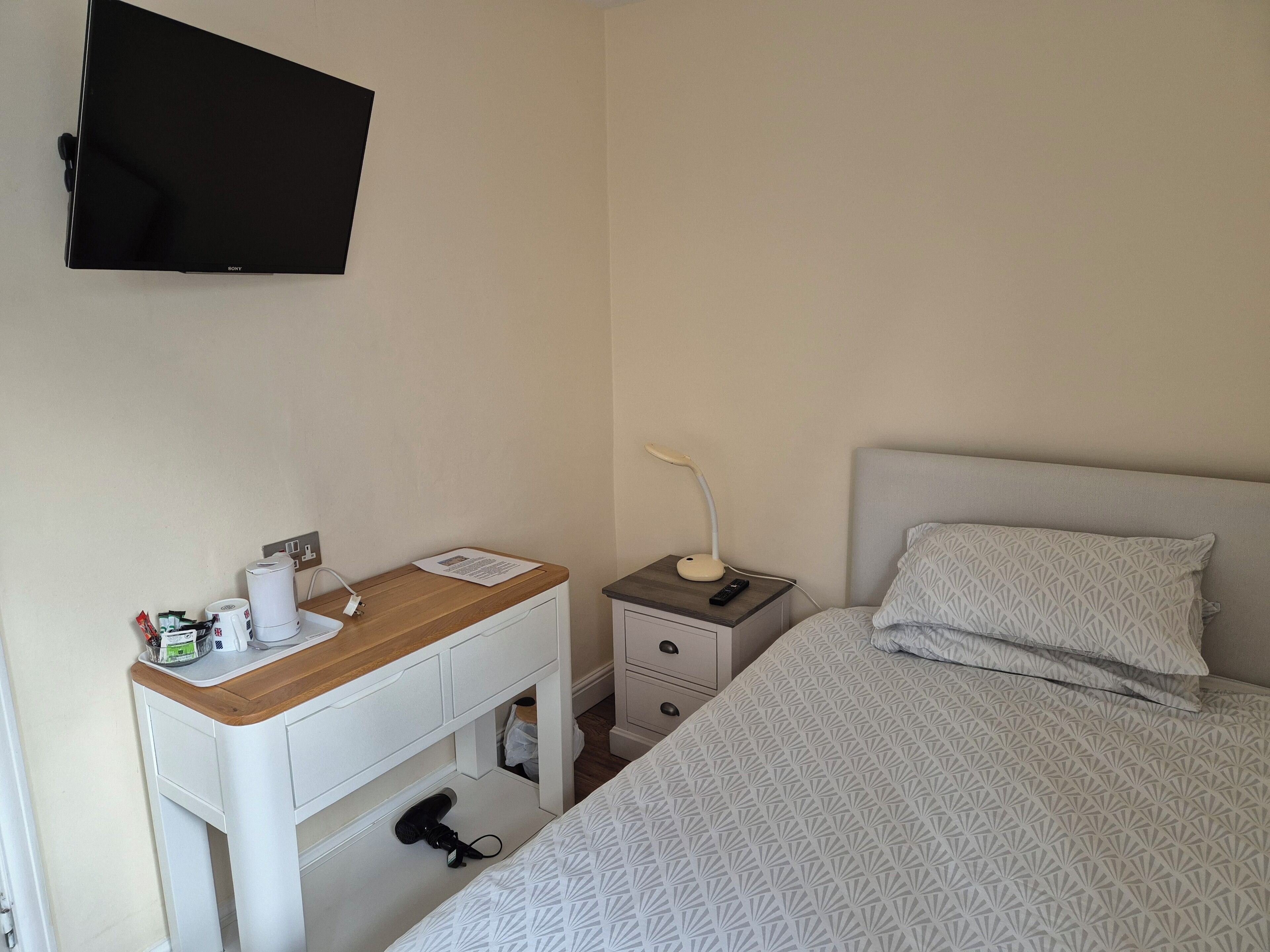Una camera da letto con un letto, un comodino con una lampada, una TV a parete e una scrivania con una sedia.